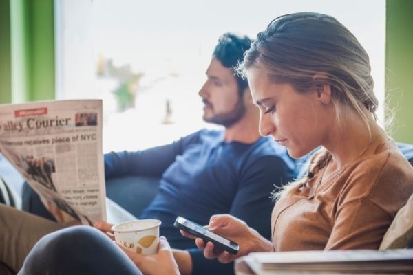 Woman looking at her phone while husband reads the newspaper.